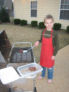 Small child in a red apron learning to cook on a grill.