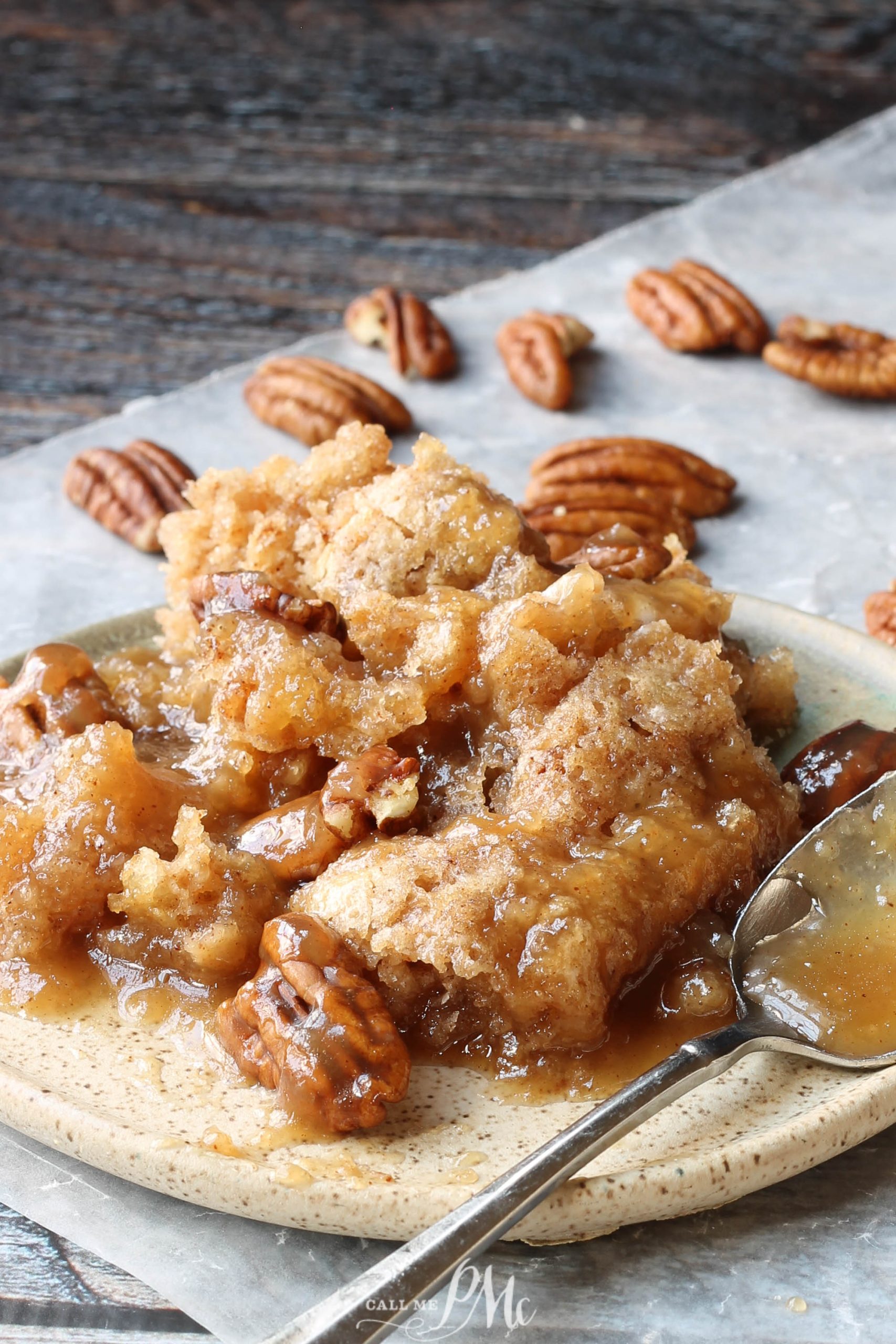 A plate of pecan pie bread pudding with caramel sauce and scattered pecans evokes the rustic charm of a pecan cobbler, all beautifully arranged on a wooden surface.