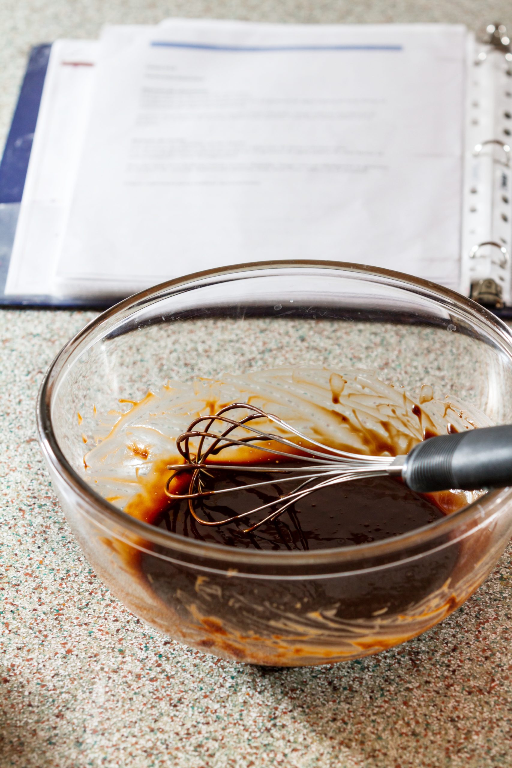 A glass bowl with brown batter and a whisk sits on the countertop, evoking a scene worthy of crafting Martha Washington Candy. An open cookbook lies in the background, promising sweet delights to come.