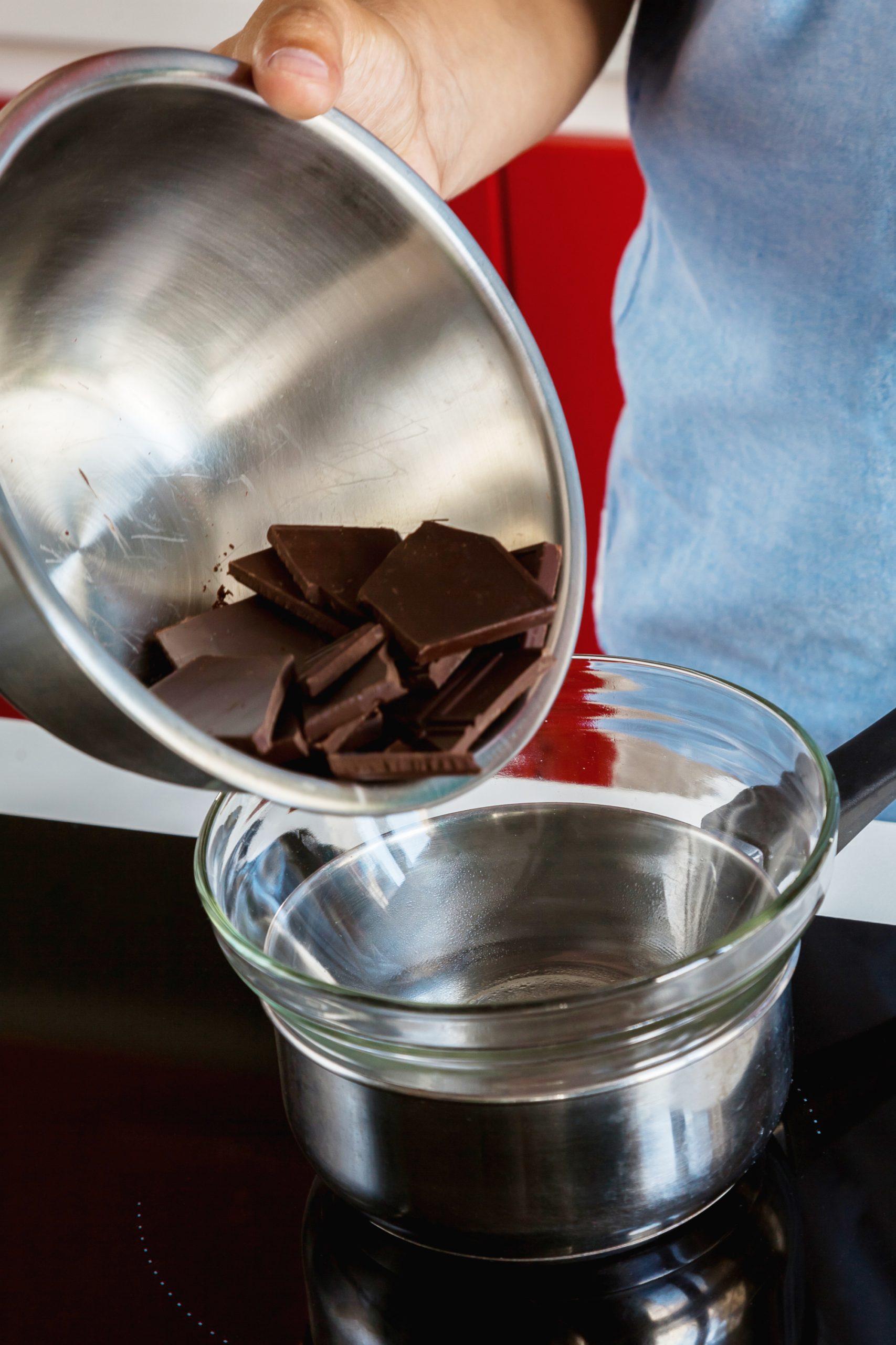 Person pouring chocolate pieces from a metal bowl into a glass bowl over a saucepan on the stove, crafting delightful Martha Washington Candy.