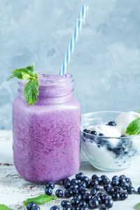 A mason jar filled with a refreshing mixed berry smoothie, complete with a blue-striped straw and garnished with mint. Next to it sits a bowl of yogurt topped with juicy blueberries, surrounded by scattered berries and sprigs of mint.