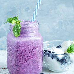 A mason jar filled with a refreshing mixed berry smoothie, complete with a blue-striped straw and garnished with mint. Next to it sits a bowl of yogurt topped with juicy blueberries, surrounded by scattered berries and sprigs of mint.