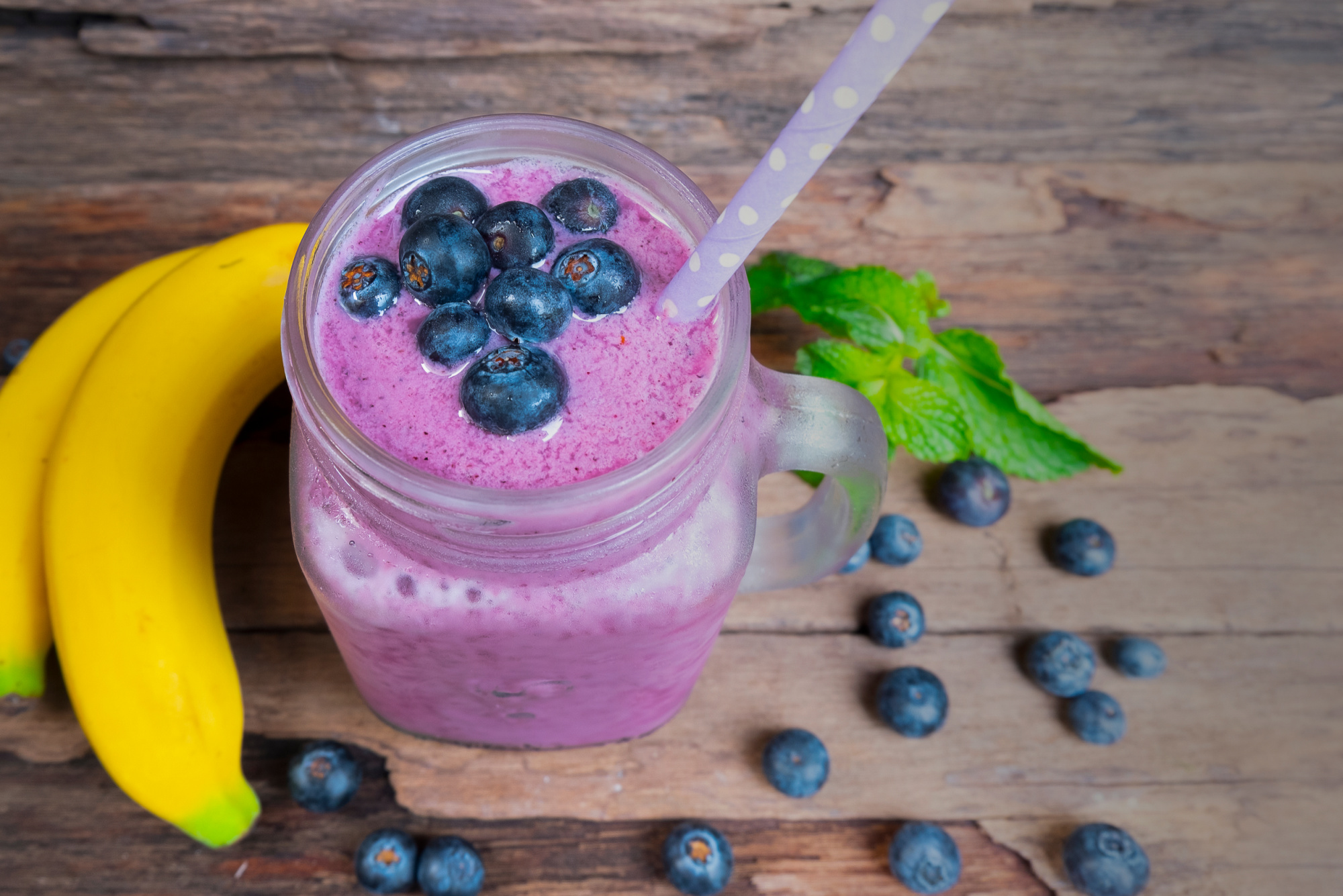 A mason jar filled with a luscious mixed berry smoothie, topped with fresh blueberries and a straw. Two bananas and scattered blueberries are beside it on a wooden surface. Mint leaves are in the background, adding a fresh touch to this delightful scene.