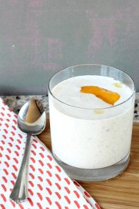 A glass of creamy Peach Greek Yogurt Smoothie with a peach slice on top sits gracefully on a wooden board, flanked by a patterned napkin and a spoon.