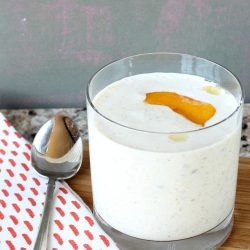 A glass of creamy Peach Greek Yogurt Smoothie with a peach slice on top sits gracefully on a wooden board, flanked by a patterned napkin and a spoon.