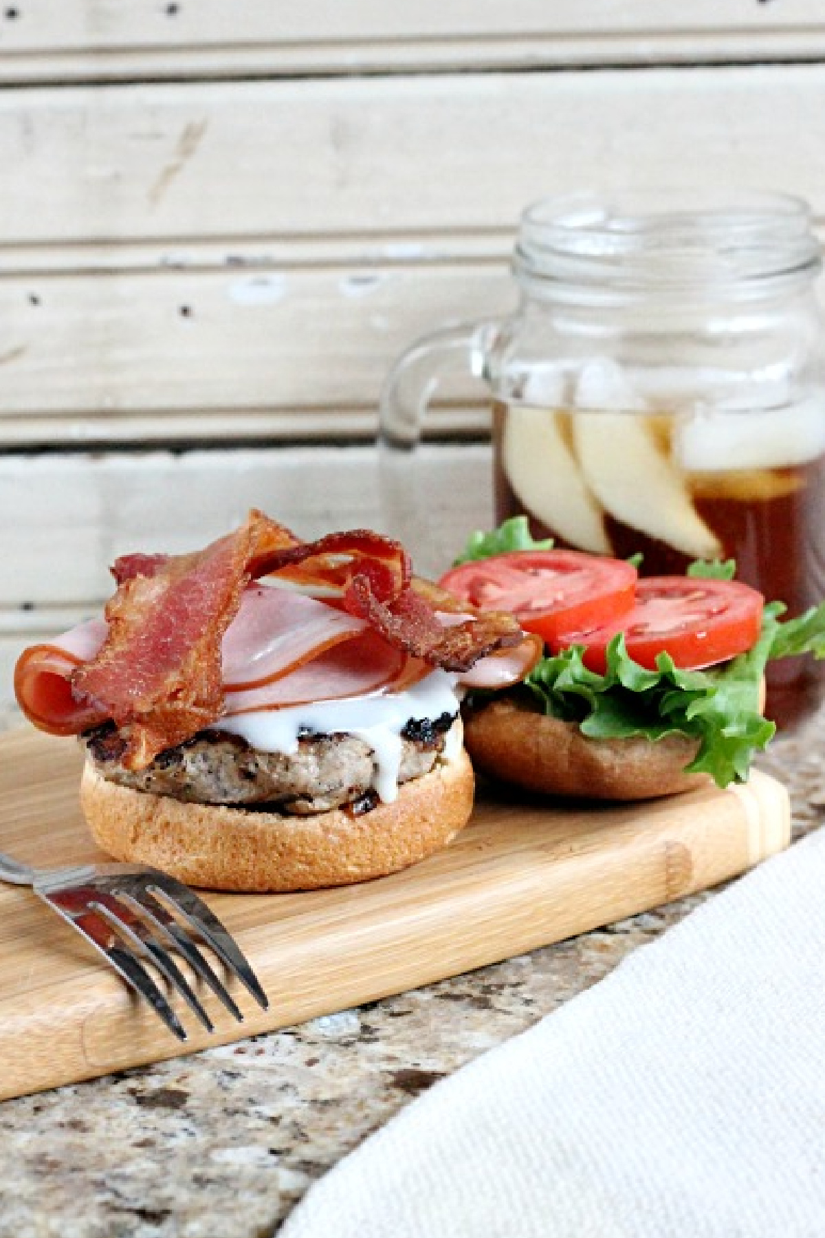 The Kentucky Hot Brown Burger boasts bacon, meat, and cheese on a wooden board, accompanied by a bun with lettuce and tomato slices. A refreshing glass of iced tea accents the scene in the background.