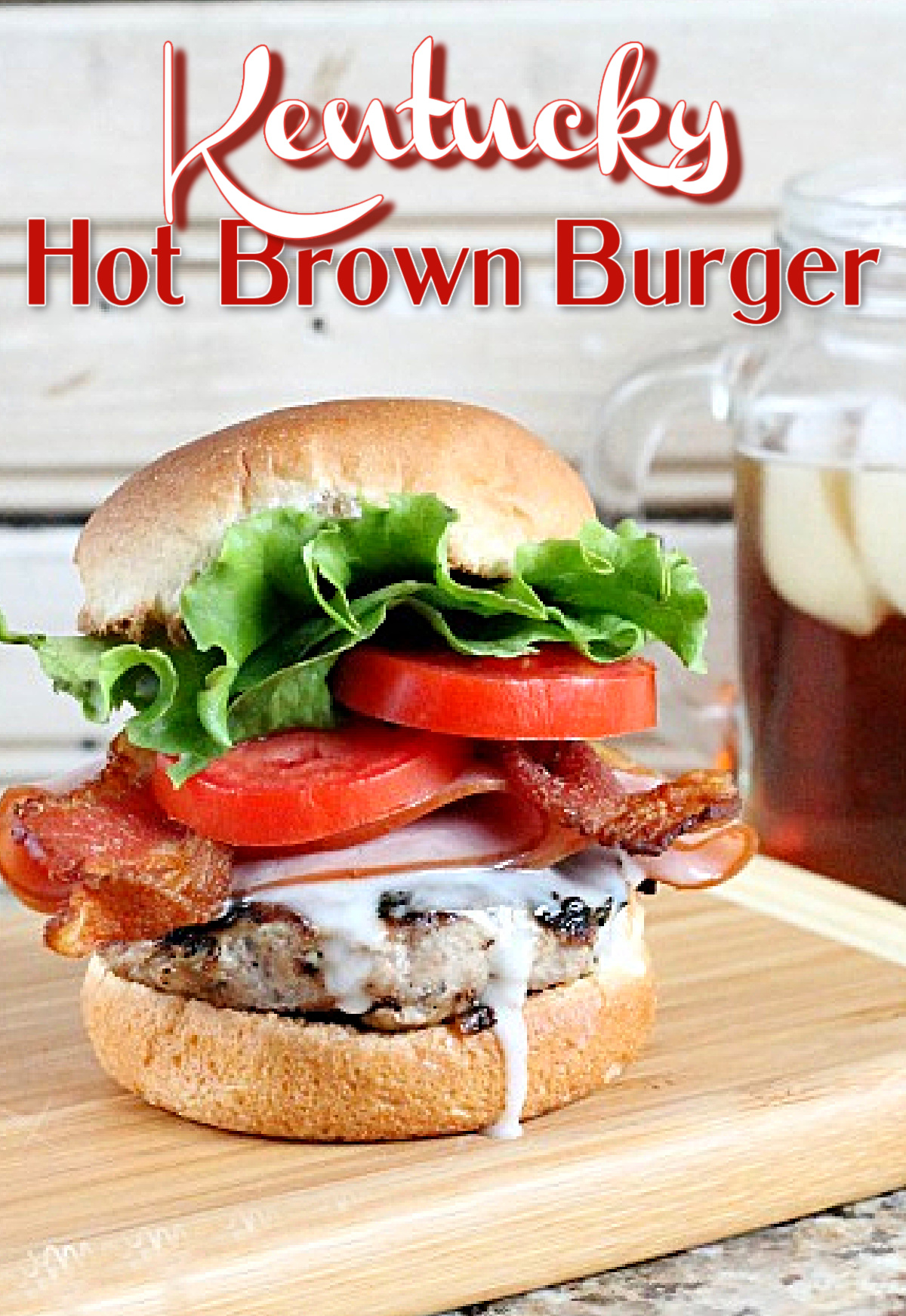 A delectable Kentucky Hot Brown Burger, topped with crisp lettuce, juicy tomato, savory bacon, and creamy white sauce, rests invitingly on a wooden board. A refreshing iced drink awaits nearby.