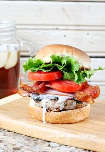 A Kentucky Hot Brown Burger with bacon, lettuce, tomato, and savory sauce sits enticingly on a wooden board. Nearby, a jar of refreshing iced tea completes this delightful scene.