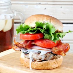 A Kentucky Hot Brown Burger with bacon, lettuce, tomato, and savory sauce sits enticingly on a wooden board. Nearby, a jar of refreshing iced tea completes this delightful scene.