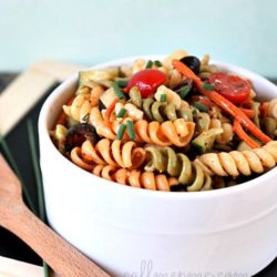 A white bowl brimming with vibrant vegetable pasta salad features tri-color rotini, garnished with cherry tomatoes, olives, and herbs. It rests on a black surface beside a wooden spoon, ready to entice your taste buds.