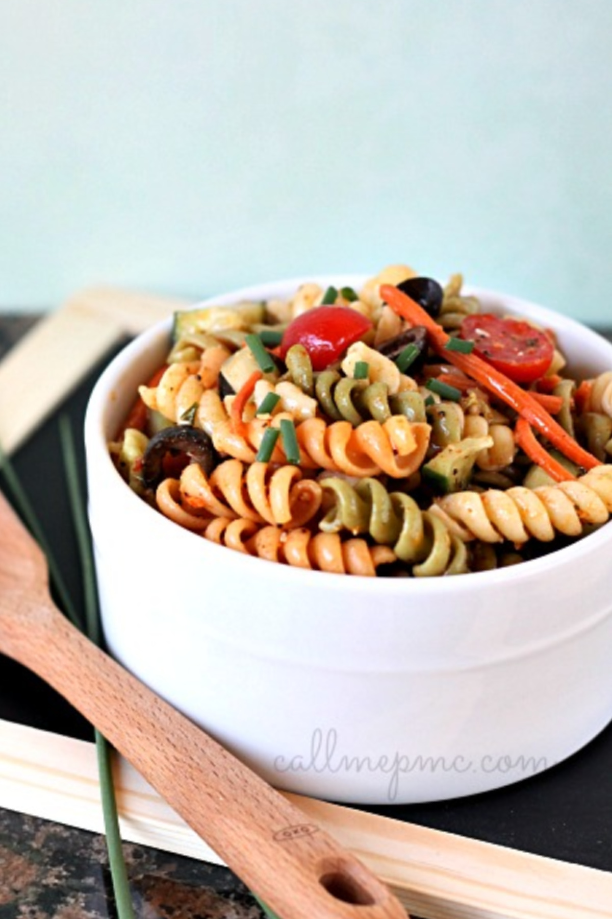 A white bowl brimming with vibrant vegetable pasta salad features tri-color rotini, garnished with cherry tomatoes, olives, and herbs. It rests on a black surface beside a wooden spoon, ready to entice your taste buds.