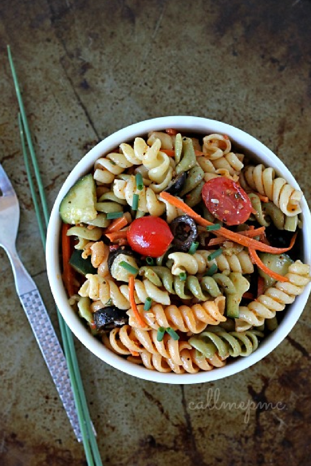 A bowl of vibrant vegetable pasta salad featuring tri-color rotini, cherry tomatoes, cucumber, carrots, and fresh herbs. A fork and chive stalks rest beside the dish.