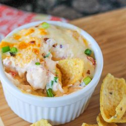 A white ramekin brimming with Hot Crawfish Cheese Dip, topped with melted cheese and green onions, sits surrounded by corn chips on a wooden surface.