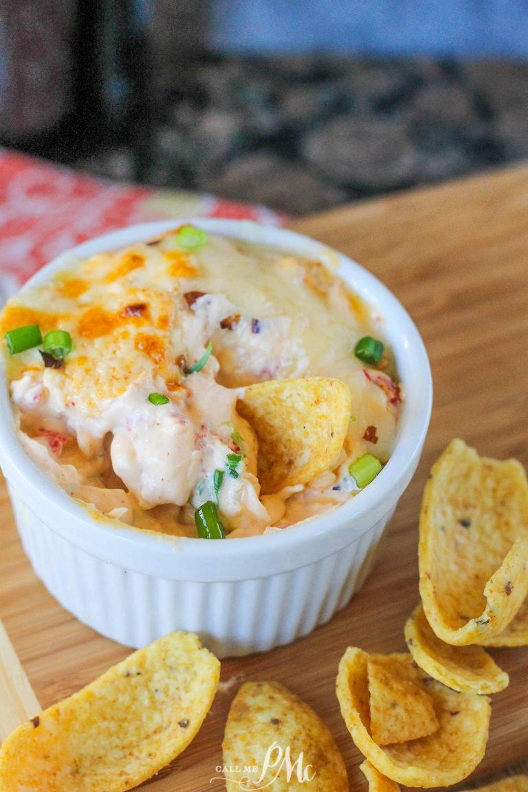 A white ramekin brimming with Hot Crawfish Cheese Dip, topped with melted cheese and green onions, sits surrounded by corn chips on a wooden surface.