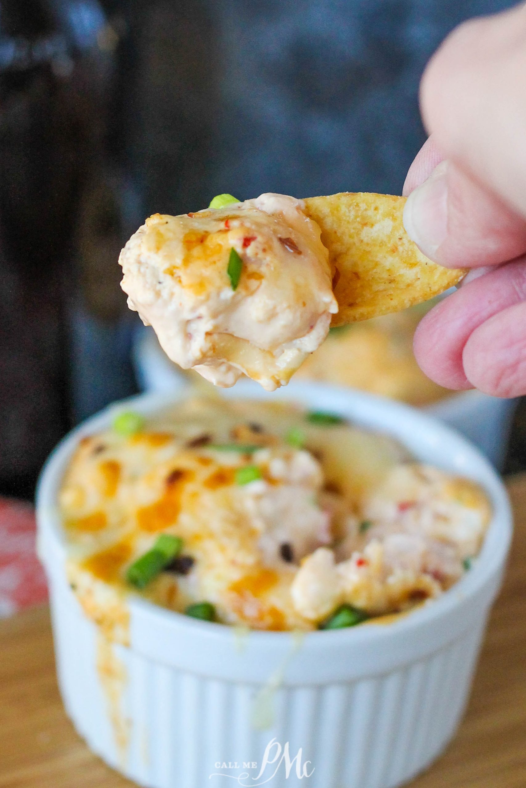 A hand holds a chip with dip, topped with herbs, over a ramekin filled with the same creamy concoction.