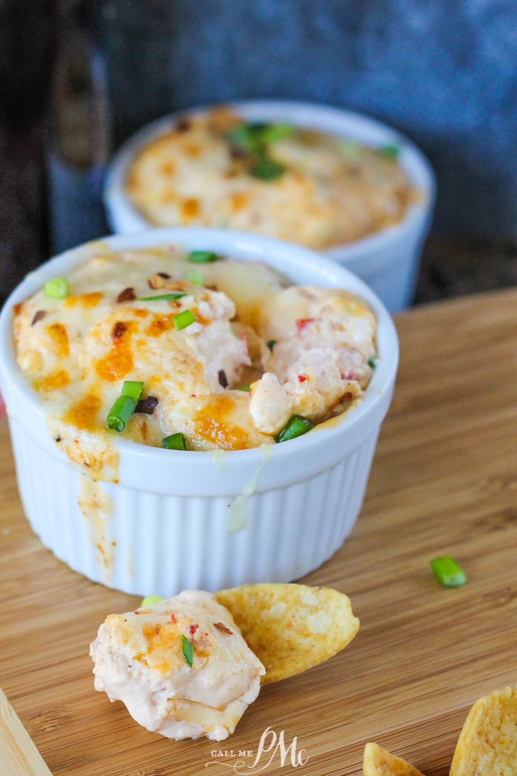 Two ramekins of hot crawfish cheese dip topped with melted cheese and chopped green onions, served alongside crispy tortilla chips on a wooden board.