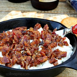 A skillet brimming with creamy corn bacon dip and savory bacon pieces rests on a wooden table, accompanied by crackers and two chilled bottles of beer in the background.