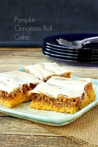 Four pieces of delectable Pumpkin Cinnamon Roll Cake with frosting rest on a rectangular plate, placed on a wooden surface with a stack of black plates and metal forks in the background.