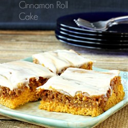 Four pieces of delectable Pumpkin Cinnamon Roll Cake with frosting rest on a rectangular plate, placed on a wooden surface with a stack of black plates and metal forks in the background.