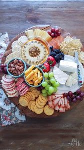 Learn how to assemble a charcuterie platter featuring an array of assorted cheeses, crackers, sliced meats, grapes, strawberries, nuts, hummus, and blackberries elegantly arranged on a rustic wooden tray.