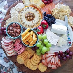 Learn how to assemble a charcuterie platter featuring an array of assorted cheeses, crackers, sliced meats, grapes, strawberries, nuts, hummus, and blackberries elegantly arranged on a rustic wooden tray.