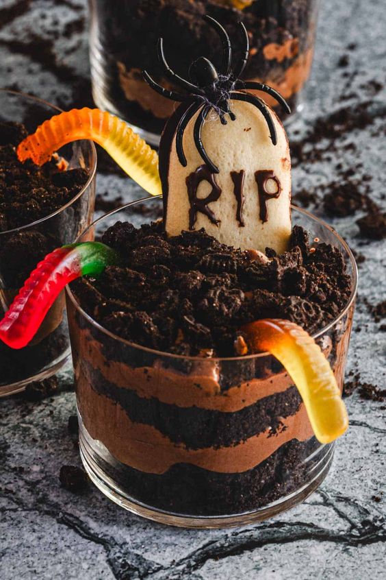 A creative and creepy Halloween dessert features layers of chocolate and cookie in a glass, topped with a "RIP" cookie, gummy worms, and a plastic spider, resembling a miniature graveyard scene.