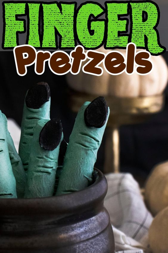 Close-up of green, finger-shaped pretzels with black fingernails in a black pot, labeled "Finger Pretzels." This creative Halloween food sits in front of pumpkins and a dark cloth, adding a creepy touch to the scene.