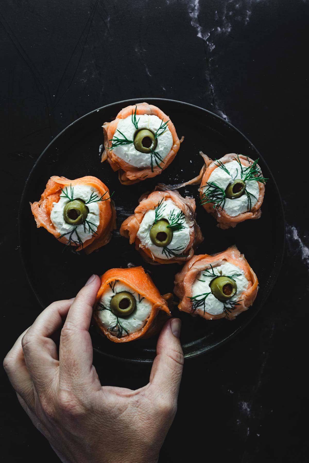 A hand reaching for a plate of six smoked salmon rolls garnished with cream cheese, green olives, and dill, arranged on a black surface. This creatively creepy Halloween food is sure to delight and spook your guests!