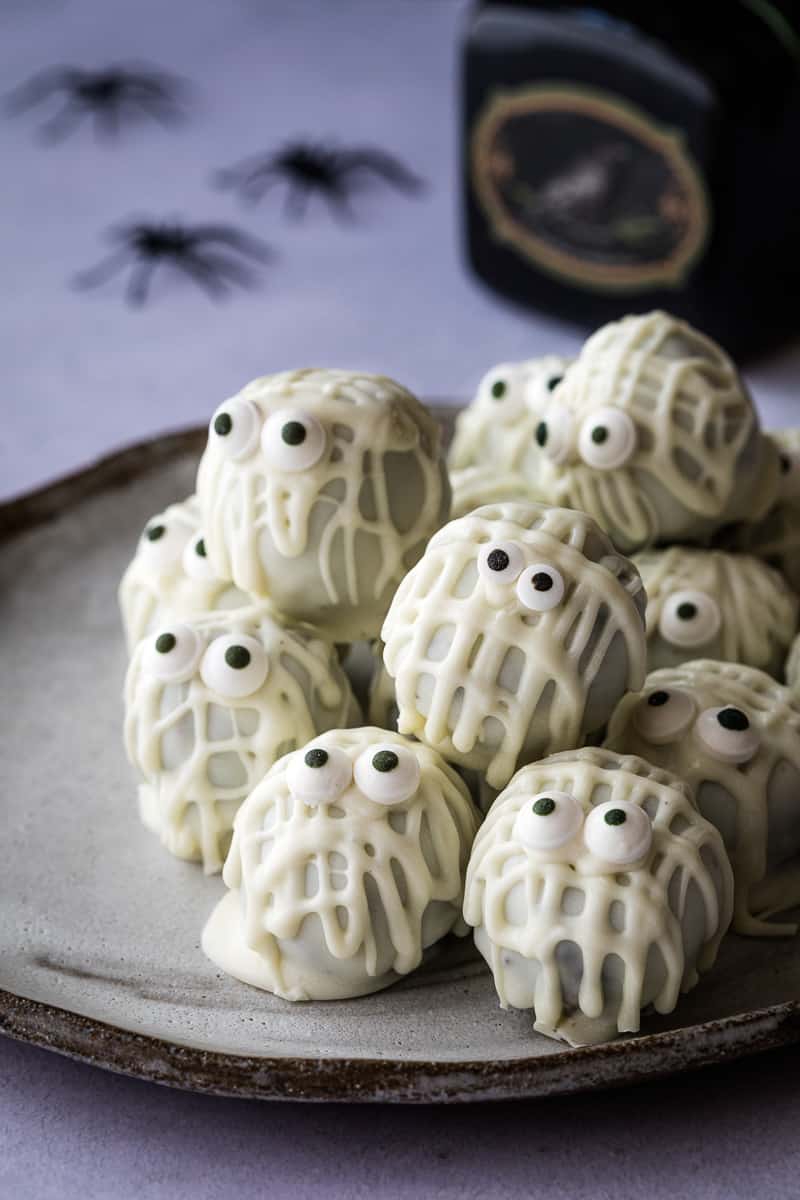 A plate of white chocolate-coated treats decorated with icing and candy eyes to resemble mummies, with small plastic spider decorations in the background, makes for some truly creative Halloween food.