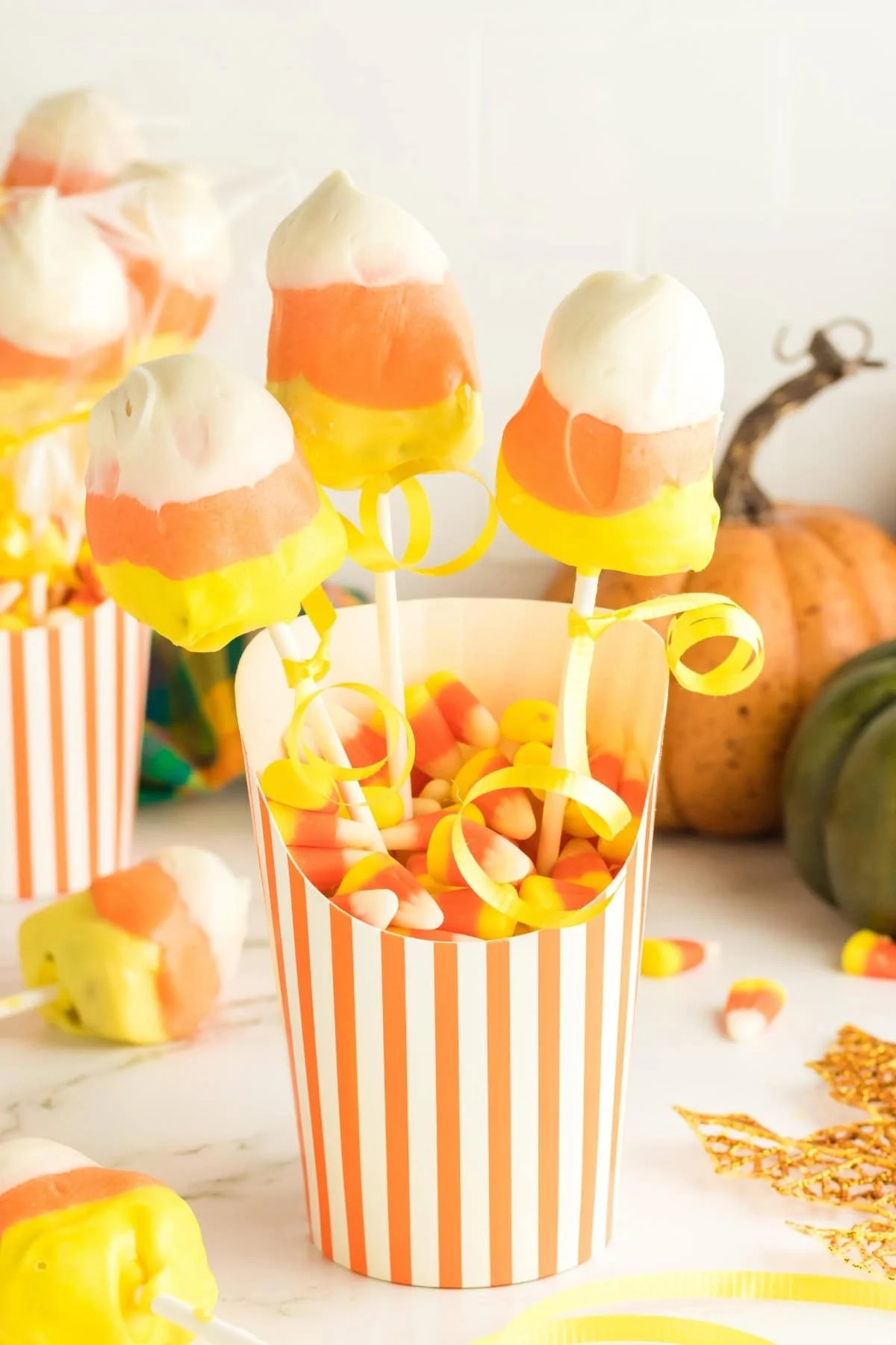 Candy corn marshmallow pops in a striped cup, surrounded by colorful decorations, create a festive and creative Halloween treat with candy corn and a pumpkin.