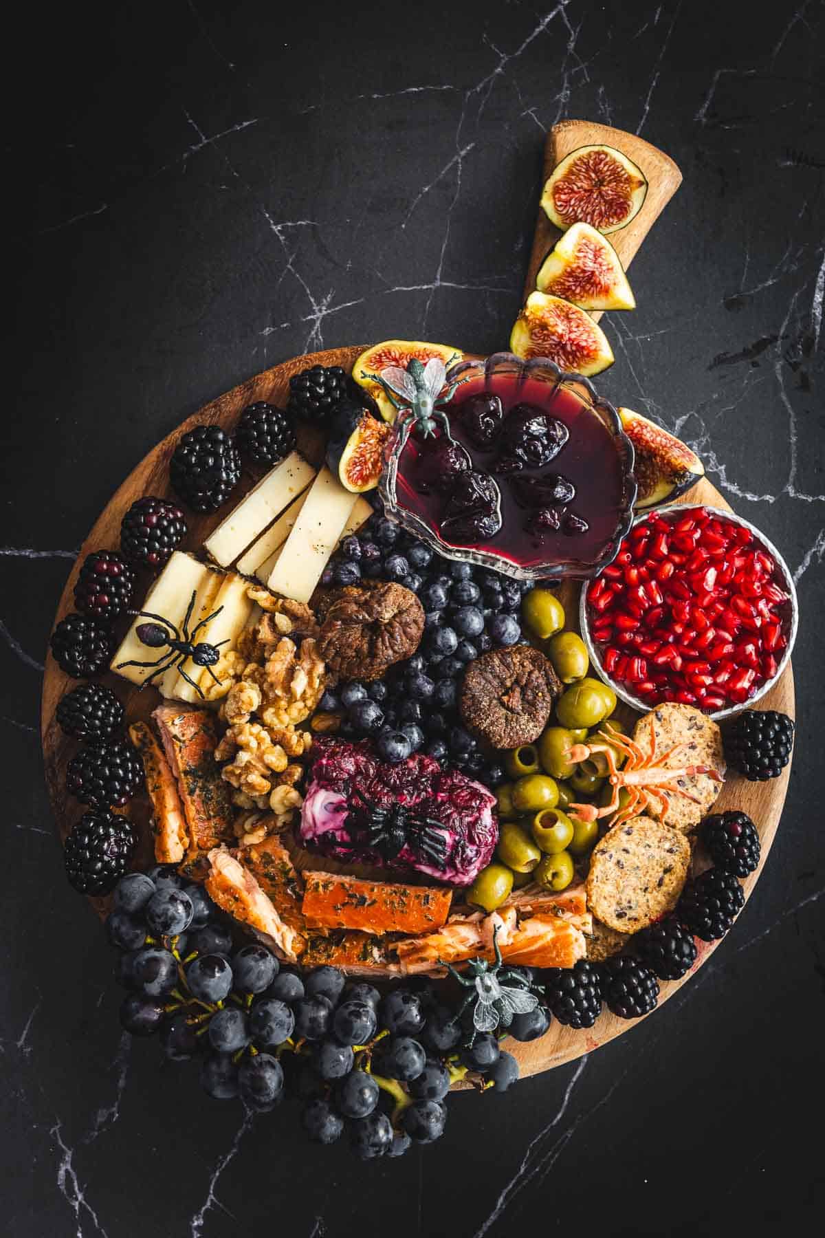 A wooden board is filled with various cheeses, berries, figs, pomegranate seeds, olives, nuts, and crackers. A small bowl of jam is placed on top. This creative spread sits on a dark surface—perfect for a slightly creepy Halloween feast.