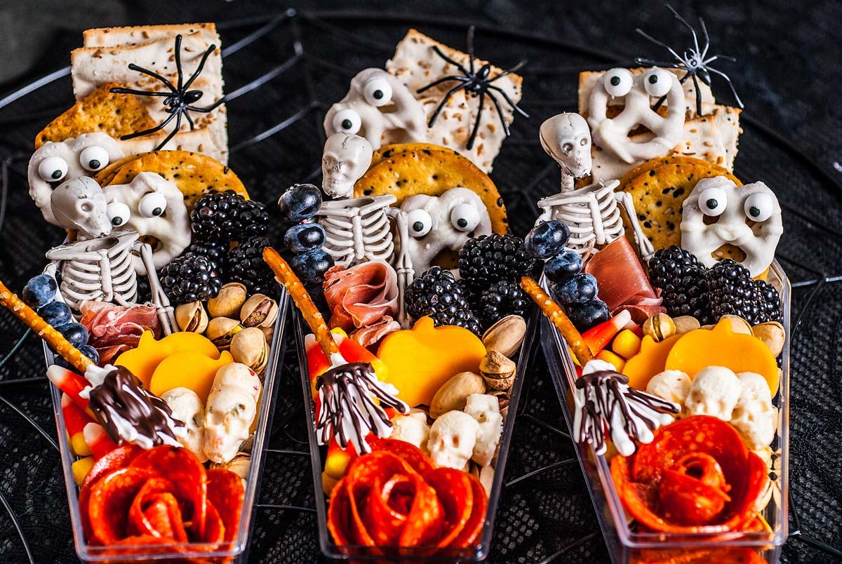 Three creative Halloween-themed snack trays featuring crackers, cheeses, meats, berries, pretzels, nuts, and creepy decorations of skeletons, ghosts, and spiders.