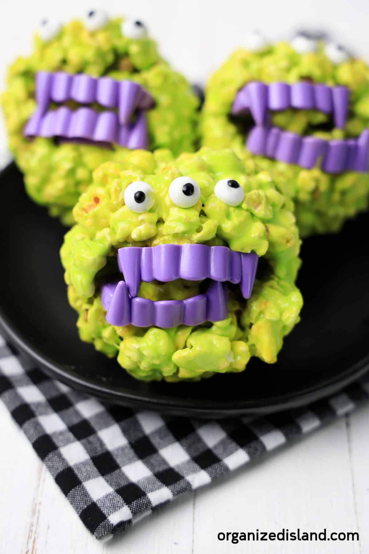 Three creative, green monster-themed Halloween treats with purple candy teeth and googly eyes, displayed on a black plate with a black-and-white checkered cloth.