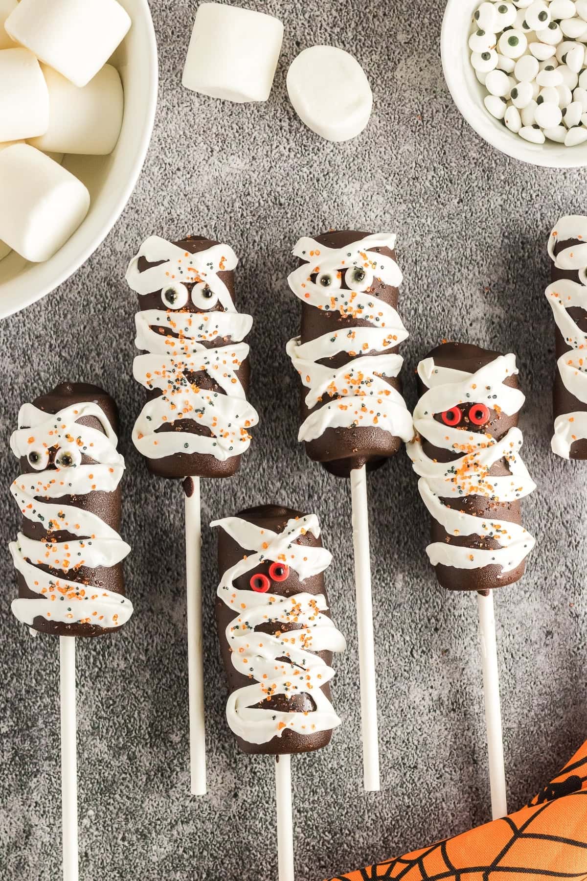 Chocolate cake pops decorated to look like mummies with white frosting and candy eyes are arranged on a gray surface next to a bowl of marshmallows and candy eyeballs, offering a creative take on creepy Halloween food.