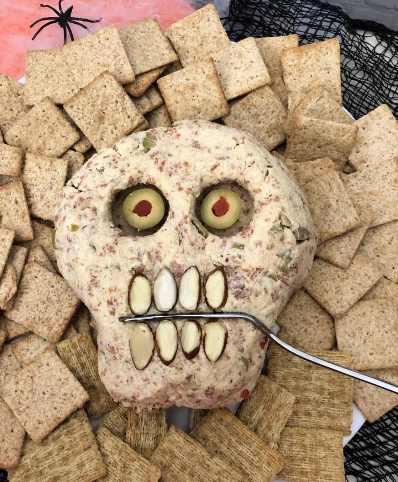 A creatively creepy Halloween-themed cheese ball shaped like a skull, featuring almonds for teeth and olives for eyes, is surrounded by crackers with a spoon resting in the mouth area.