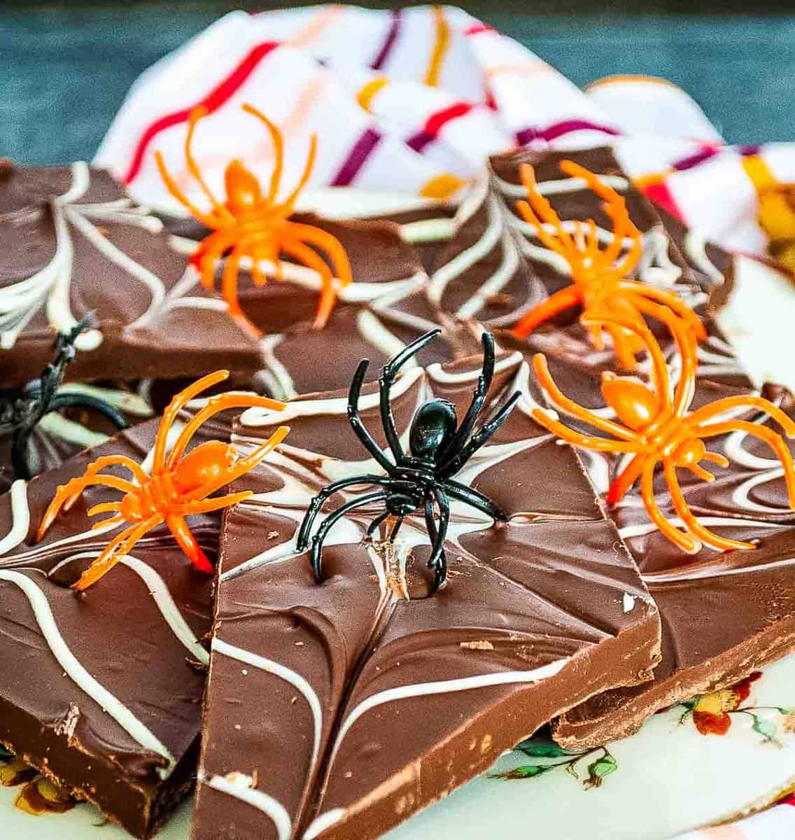 A plate of chocolate bark, creatively adorned with white chocolate swirls and topped with plastic spiders, rests on a patterned cloth—an eerie yet delightful Halloween treat.