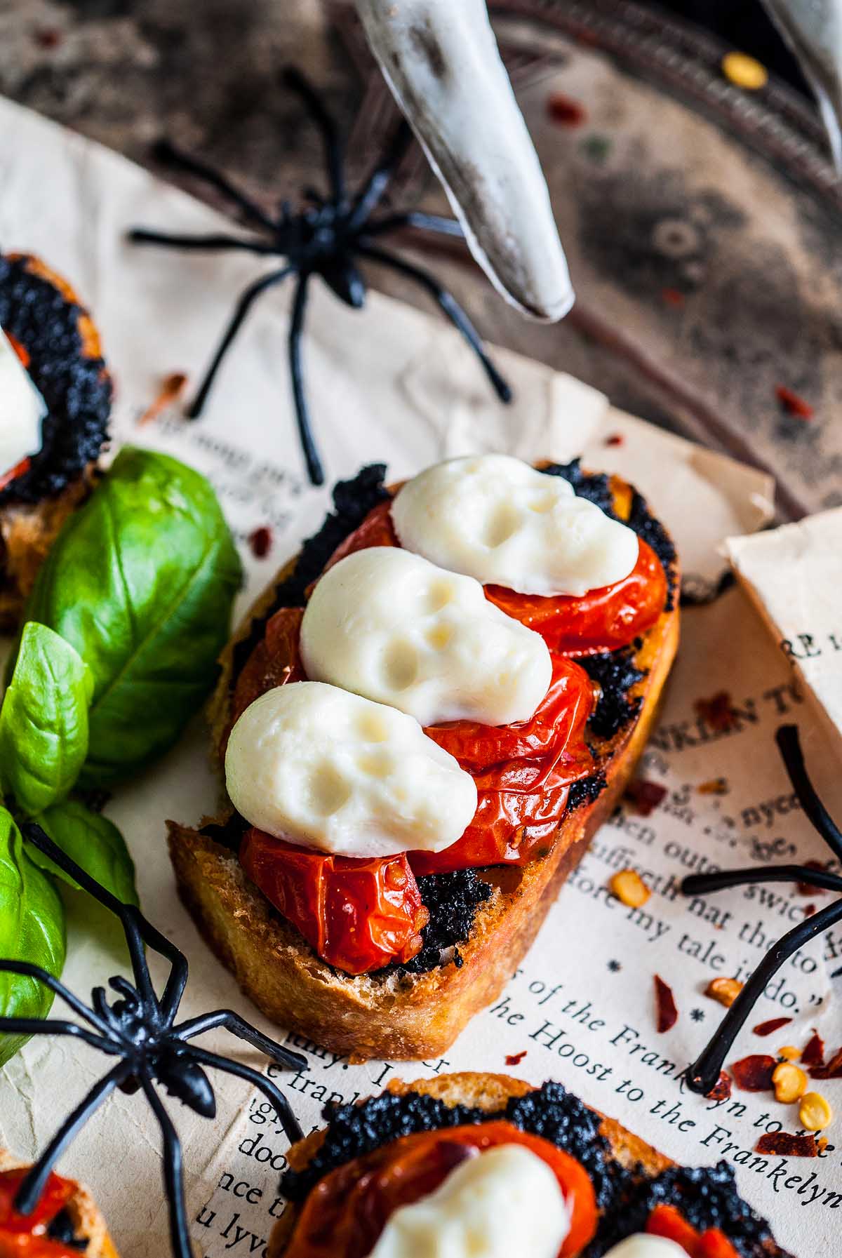 A slice of toast topped with black spread, cherry tomatoes, and mozzarella shaped like skulls, surrounded by fake spiders and basil leaves—perfectly creepy Halloween food for a creative twist.