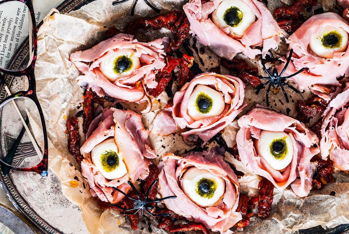 A tray of creative, Halloween-themed snacks made from ham slices with a center resembling an eye, garnished with black olives and mozzarella cheese, surrounded by fake spiders and dried tomatoes. Enjoy some creepy fun this season!