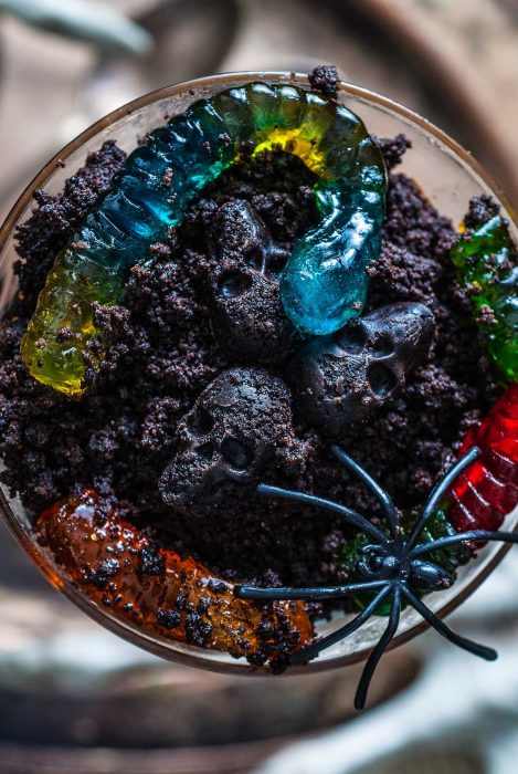 A creative Halloween dessert with crushed cookies resembling dirt, topped with gummy worms, black gummy skulls, and a plastic spider for a creepy twist.