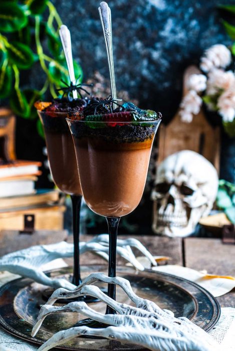 Two dark-stemmed glasses filled with chocolate mousse, topped with gummy worms and a cookie crumble, are placed on a silver platter with decorative skeletal hands. This creative yet creepy Halloween food is perfectly complemented by a skull and greenery in the background.