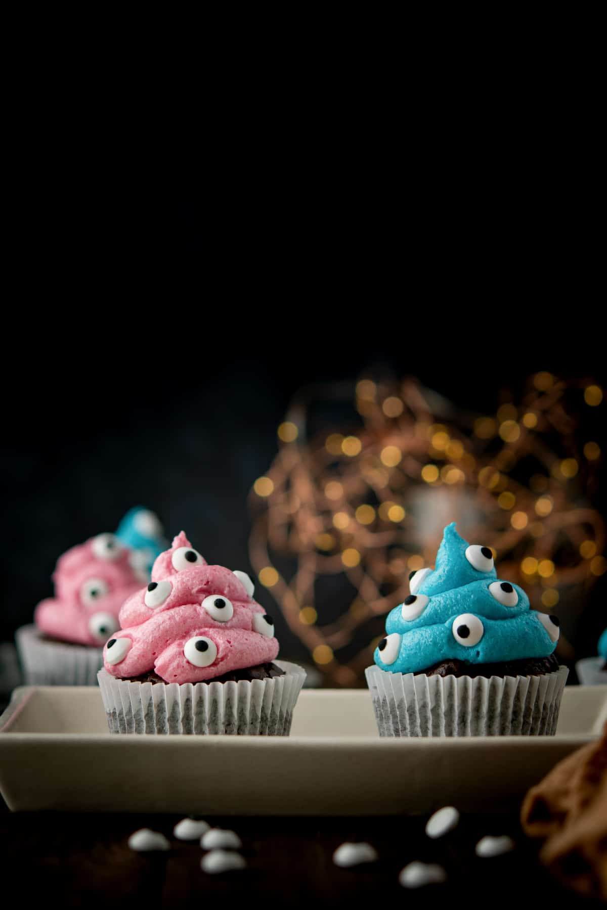 Cupcakes with pink and blue frosting, decorated with candy eyes, are placed on a rectangular tray. A blurred string of lights is visible in the background, adding to the creative yet slightly creepy Halloween vibe.