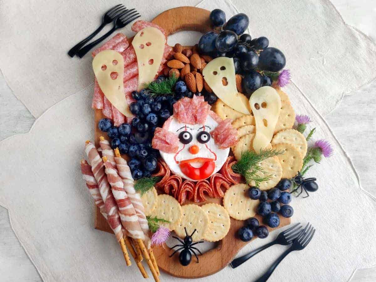 A wooden board displays a creative creepy Halloween-themed charcuterie arrangement with crackers, cheese slices shaped like ghosts, grapes, blueberries, almonds, salami, and a cheese ball decorated as a clown face.