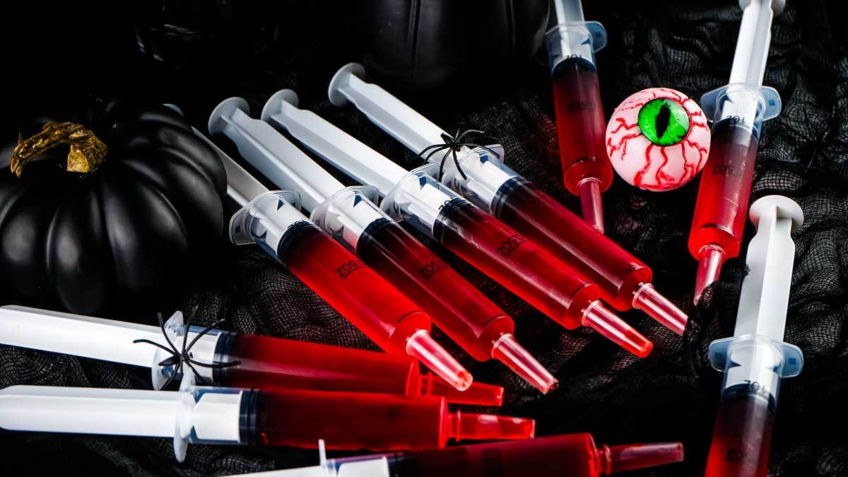 Several syringes filled with red liquid are scattered on a black fabric, accompanied by a plastic eyeball and a black pumpkin, creating a creepy Halloween scene that sparks creative ideas for themed food.