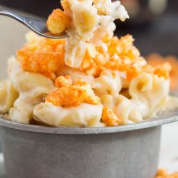 A fork holds a bite of Eight Cheese Mac n Cheese, topped with crushed Cheetos, above a metal cup filled with more of the cheesy delight.