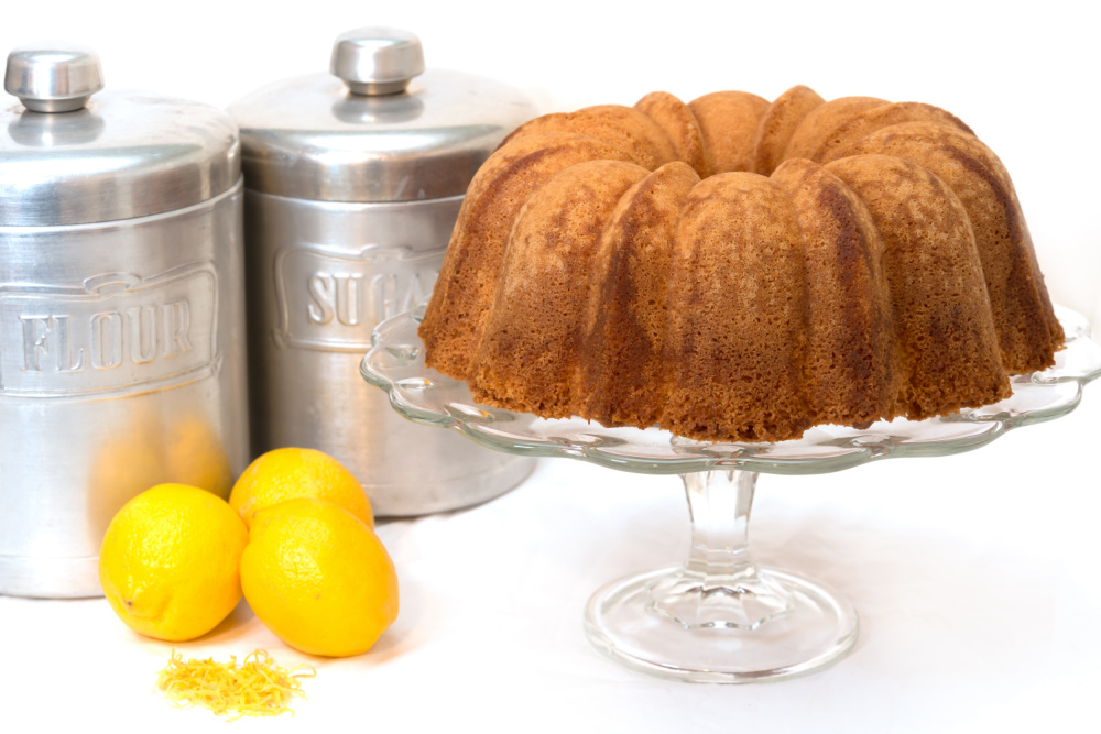 A bundt cake on a glass stand, with two metal canisters labeled "FLOUR" and "SUGAR" in the background. Three lemons and lemon zest are placed beside the stand, adding a zesty twist akin to churro cubes.