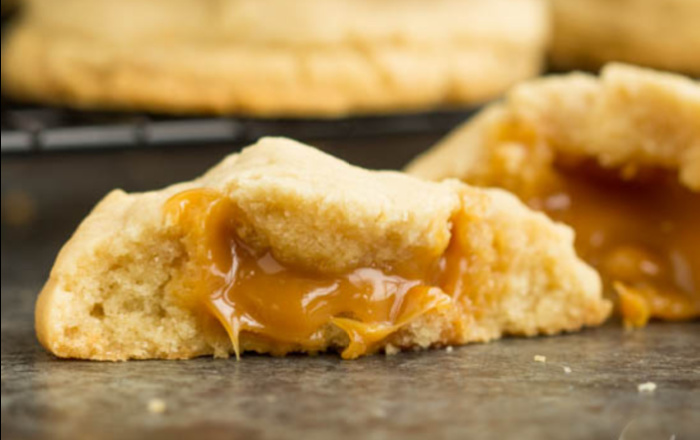 A gooey caramel-stuffed sugar cookie, partially broken open, rests on a dark surface.