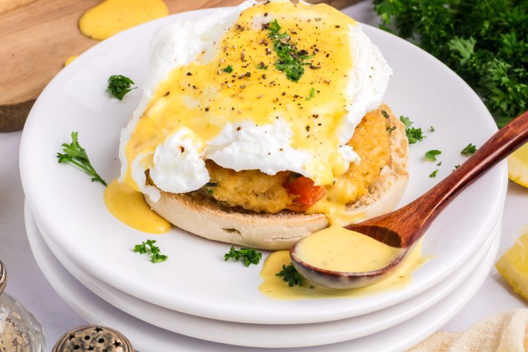 A plate with an English muffin crowned by a poached egg, drizzled with a velvety hollandaise sauce made using a blender, and sprinkled with herbs. A wooden spoon rests beside it, ready to scoop up extra sauce.