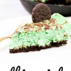 A slice of Thin Mint Cheesecake on a white plate with a fork, boasting a chocolate crust and mint-flavored filling. A full cheesecake is partially visible in the background.