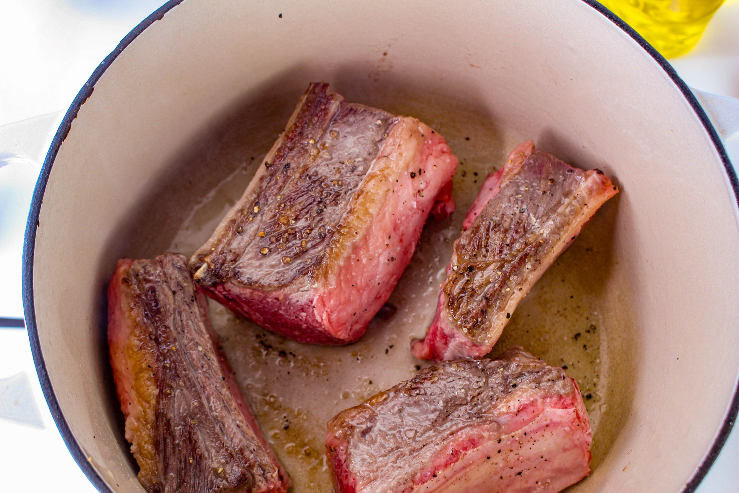 Four pieces of browned meat in a white pot, partially cooked and seasoned with pepper, evoke the rich aroma of balsamic braised short ribs.