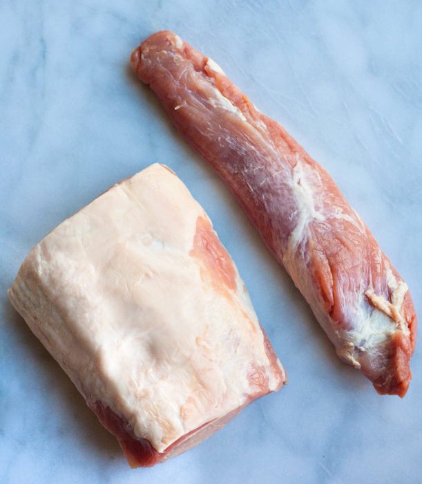 Two raw cuts of pork on a white marble surface: one rectangular piece with visible fat, perfect for transforming into crispy bacon, and one elongated, lean tenderloin.